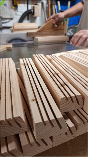 Workshop view of wooden playing card holders being made in batches, with unfinished fir holders stacked in front and the maker working at a table saw.