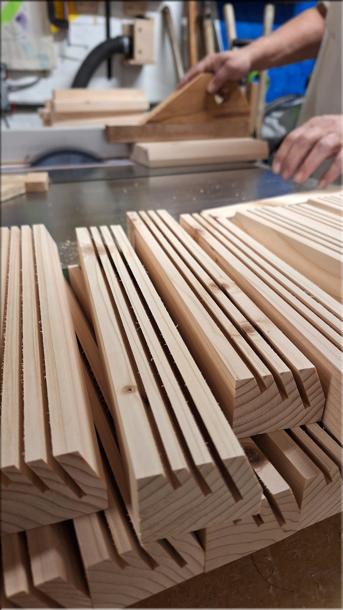 Workshop view of wooden playing card holders being made in batches, with unfinished fir holders stacked in front and the maker working at a table saw.